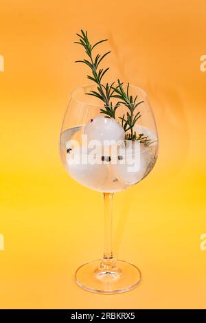 Studioaufnahme eines Gin Tonic Cocktails mit Rosmarin, Pfeffer und Eiswürfeln, isoliert in Glas vor frischem gelben Hintergrund Stockfoto