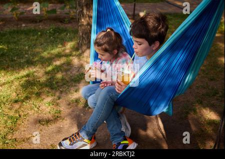 Zwei verschiedene Kinder, ein weißer Teenager und ein Vorschulmädchen in lässiger Kleidung, trinken Saft aus Stroh, sitzen in einer Hängematte, entspannen zusammen in einem Stockfoto