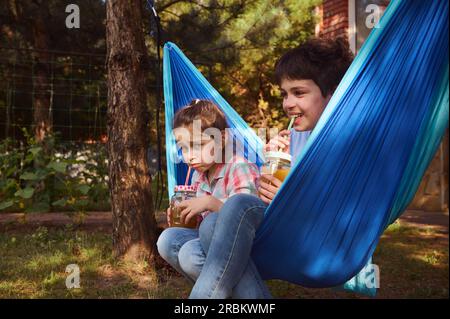 Glückliche Kinder im Grundalter haben Spaß zusammen, lächeln, entspannen in der blauen Hängematte im Sommerlager, trinken gesunden Smoothie aus Stroh. Kinder. Childho Stockfoto