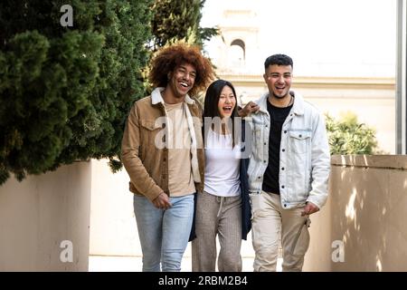 Drei fröhliche junge Menschen lächeln und sehen entspannt aus, wenn sie auf der Straße spazieren gehen. Eine multiethnische Gruppe, die Spaß hat und sich gegenseitig umarmt, während sie im Park spazieren. Stockfoto