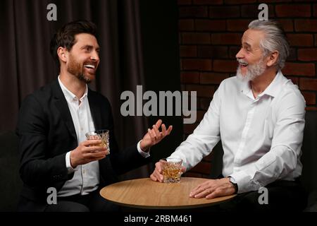 Männer mit Whiskey-Gläsern, die sich drinnen an einem Holztisch unterhalten Stockfoto