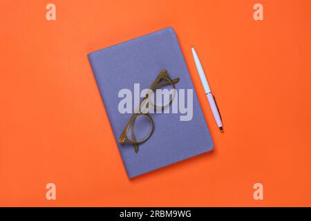 Geschlossenes blaues Notizbuch, Brille und Stift auf orangefarbenem Hintergrund, flach liegend Stockfoto