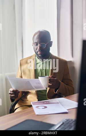 Junger seriöser Geschäftsmann mit einer Tasse Kaffee, der Finanzdokumente durchsucht, während er am Arbeitsplatz vor der Kamera im Büro sitzt Stockfoto