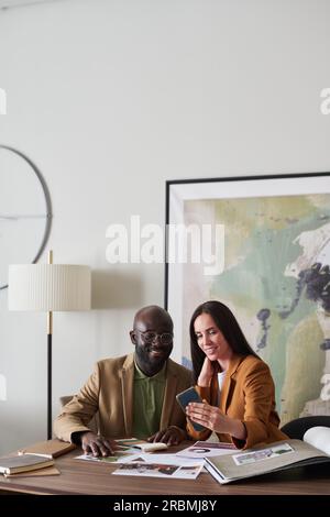 Eine junge lächelnde Innendesignerin, die einem männlichen Kollegen Online-Skizzen oder Fotos zeigt, während sie über neugierige Skizzen diskutiert und sie auswählt Stockfoto