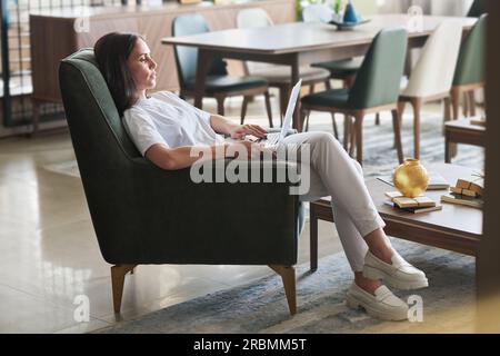 Junge, braune Frau in legerer Kleidung, die ihren Laptop benutzt, während sie im Wohnzimmer in einem bequemen grauen Samtsessel sitzt und im Netz arbeitet Stockfoto