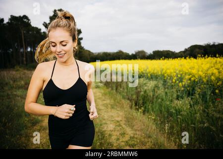 Junge Laufsportlerin, die auf einer felsigen Landstraße neben einem Blumenfeld arbeitet eine junge Frau in Sportbekleidung, die auf einer Straße läuft. Stockfoto