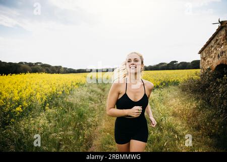 Junge Laufsportlerin, die auf einer felsigen Landstraße neben einem Blumenfeld arbeitet eine junge Frau in Sportbekleidung, die auf einer Straße läuft. Stockfoto