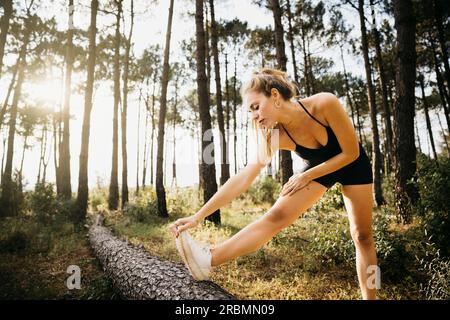 Junge Läuferin, die sich auf einem Baumstamm streckt. Fitness-Frau, die eine Pause vom Training im Freien in einem Wald macht. Stockfoto