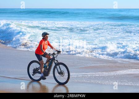 Aktive Frau, die ihr elektrisches Mountainbike am Strand von Cape Trafalgar, Costa de la Luz, Andalusien, Spanien, fährt Stockfoto