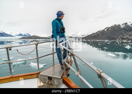 Reifer weißer Mann, der auf der Bugplattform eines Boots steht und eine Szene mit Eisberg im Wasser beobachtet, Columbia Glacier; Prince William Sound, Alask Stockfoto