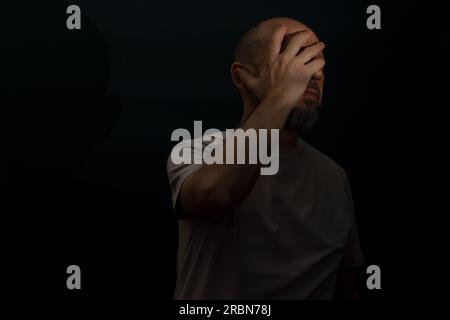 Trauriger Mann mit Kopfschmerzen oder Migräne. Gestresster Kerl mit schmerzhaftem Gesichtsausdruck, der sich schrecklich schwach oder depressiv fühlt. Isoliert auf dunklem Bac Stockfoto