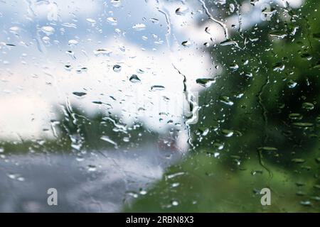Regentropfen auf der Windschutzscheibe mit Nahaufnahme des Himmels und grünen Bäumen, die im Hintergrund verschwommen sind. Fahrt auf einer im Frühling regnerischen Landstraße. Stockfoto
