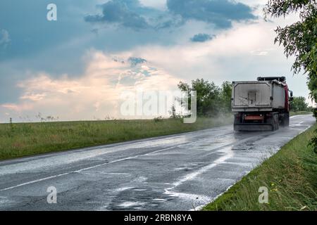 Schweres Lkw-Fahrzeug auf nassen Asphaltstraßen bei stürmischem Regen mit epischen Wolken und grüner Landschaft. Rückfahransicht mit Wassernebel auf w Stockfoto