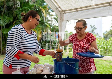 Tegucigalpa, Francisco Morazan, Honduras - 28. November 2022: Old Brown Woman serviert heiße Weihnachten aus einem Blaukübel Getränk an eine andere Frau mit La Stockfoto