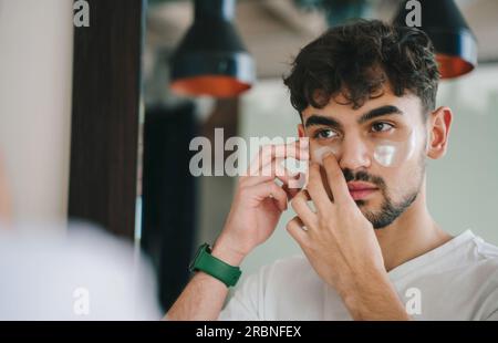 Junger bärtiger Mann in lässiger Kleidung, der morgens beim Blick in den Spiegel Pflaster aufklebt. Stockfoto