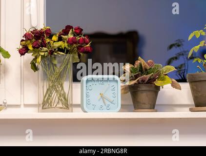 Eine gemütliche Inneneinrichtung des Hauses, Dekorationsgeräte, Uhr und Blumen vor dem Fenster, eine Gruppe von Gegenständen, Details, niemand, keine Menschen, Abend Stockfoto