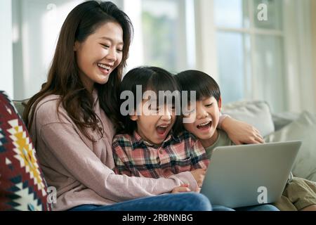 Junge asiatische Mutter und zwei Kinder, die zu Hause auf dem Familientisch sitzen und zusammen einen Laptop benutzen Stockfoto