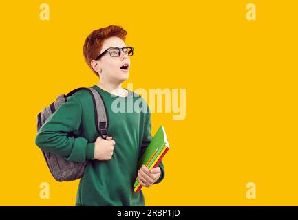 Der überraschte, schockierte, süße rothaarige Junge in transparenter Brille sieht abwesend aus, trägt einen grünen Pullover und graue Jeans, hält einen Rucksack und Lehrbücher in der Hand. Stockfoto