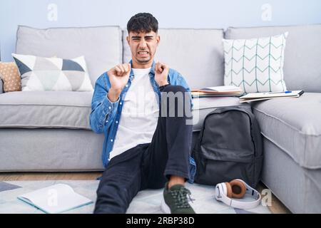 Junger hispanischer Mann, der auf dem Boden sitzt und für die Universität lernt ekelhafter Ausdruck, verärgert und ängstlich, und ekelhaftes Gesicht macht, weil Aversion reagiert Stockfoto