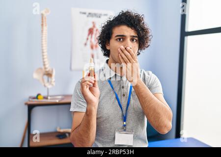 Ein hispanischer Mann mit lockigem Haar, der cbd-Öl hält, der den Mund mit der Hand bedeckt, schockiert und vor Fehlern fürchtet. Überraschter Ausdruck Stockfoto