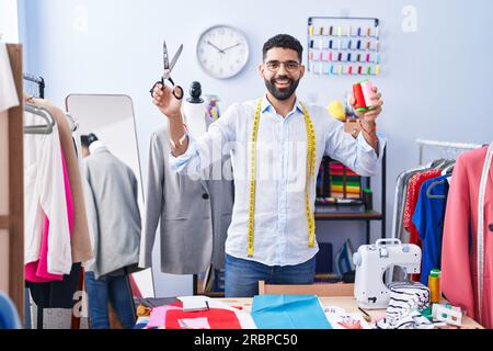 Ein junger arabischer Schneider, der selbstbewusst Schere und Faden in der Schneiderei hält Stockfoto