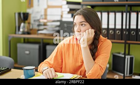 Junge, wunderschöne hispanische Geschäftsfrau, die mit ernstem Gesicht im Büro auf dem Tisch sitzt Stockfoto