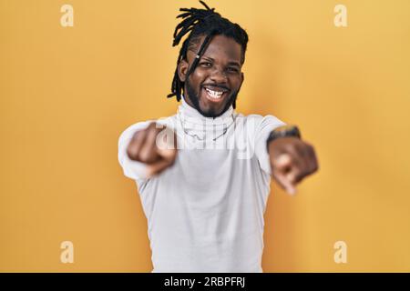 Ein afrikanischer Mann mit Dreadlocks, der einen Rollkragenpullover auf gelbem Hintergrund trägt und mit den Fingern auf dich und die Kamera zeigt, positiv lächelt und fröhlich ist Stockfoto