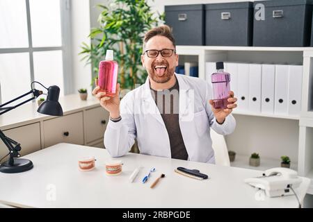 Weißer Zahnarzt mittleren Alters mit Mundspülung für frischen Atem, die Zunge mit lustigem Gesichtsausdruck ausstrecken. Stockfoto