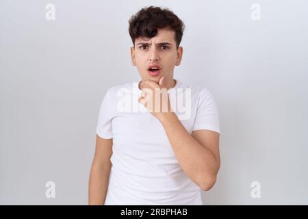 Junger nicht binärer Mann, der ein legeres weißes T-Shirt trägt, das fasziniert von Unglauben, Überraschung und erstaunlichem Ausdruck mit Händen am Kinn aussieht Stockfoto