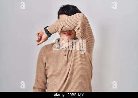 Nicht binäre Person, die über einem isolierten Hintergrund steht und die Augen mit dem Arm bedeckt, ernsthaft und traurig aussieht. Blindes, Versteckungs- und Ablehnungskonzept Stockfoto