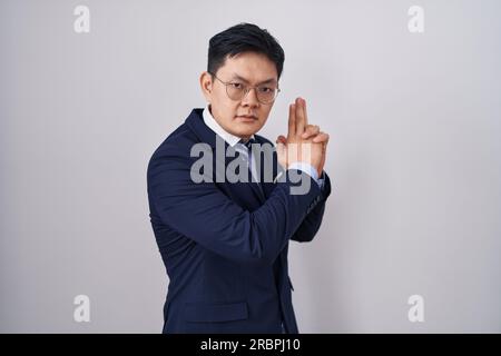 Junger asiatischer Mann in Geschäftsanzug und Krawatte, mit symbolischer Waffe, mit Handgesten, mit Tötungswaffen, wütendem Gesicht Stockfoto