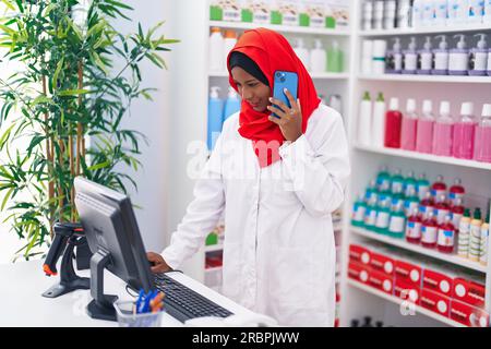 Eine junge, schöne Apothekerin, die mit einem Smartphone über einen Computer in der Apotheke spricht Stockfoto