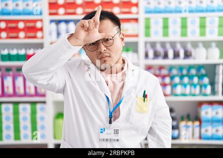 Chinesischer junger Mann, der in einer Apotheke arbeitet, macht sich über Leute mit Fingern auf der Stirn lustig, macht Verlierer-Geste, verspottet und beleidigt. Stockfoto