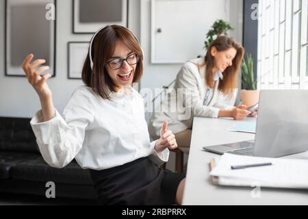 Freudige Büroangestellte mit Kopfhörern, die Musik hören und so tun, als würden sie Gitarre spielen, eine Kollegin sitzt am Schreibtisch Stockfoto