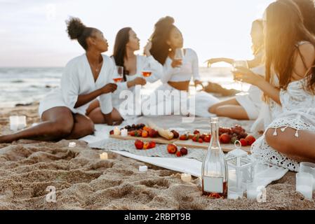 Junge, wunderschöne Frauen, die ein Sommerpicknick mit Wein und Obst am Strand bei Sonnenuntergang machen, Frauen feiern eine Hühnerparty Stockfoto