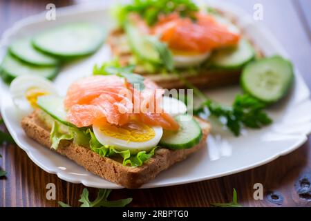 Gebratener Toast mit Salat, Ei, Gurken und rotem Fisch auf einem Teller, auf einem Holztisch. Stockfoto