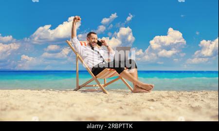 Glücklicher Geschäftsmann auf einer Liege mit Laptop und Smartphone am Meer Stockfoto