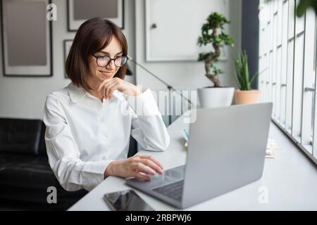 Lächelnde europäische Geschäftsfrau, die ein Notebook benutzt, online kommuniziert, E-Mails schreibt oder Kunden im Büro berät Stockfoto