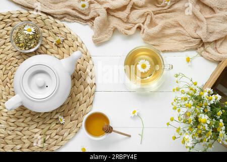 Korbmatte mit Honig, Teekanne und Tasse heißem Kamillentee auf weißem Holzhintergrund Stockfoto