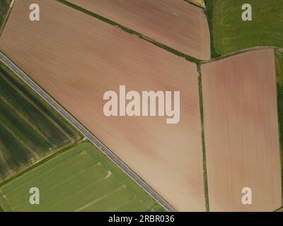 Blick auf die Drohne auf Ackerland mit braunen gepflügten Ackerfeldern, grünen Ackerfeldern und einer Asphaltstraße im Sommer Stockfoto
