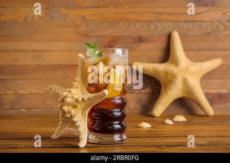 Ein Glas kalter Cuba Libre Cocktail, Seesterne und Muscheln auf Holzhintergrund Stockfoto