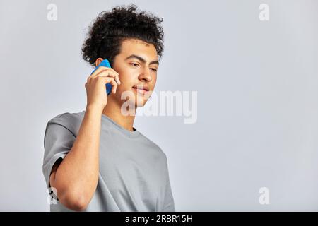 Studioporträt eines jungen, gutaussehenden brünetten Mannes mit lockigem Haar, der ein Smartphone in der Hand hält. Ein zwielichtiger Kerl lächelt und spricht mit einem Handy. M Stockfoto