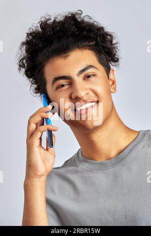 Studioporträt eines jungen, gutaussehenden brünetten Mannes mit lockigem Haar, der ein Smartphone in der Hand hält. Ein zwielichtiger Kerl lächelt und spricht mit einem Handy. M Stockfoto