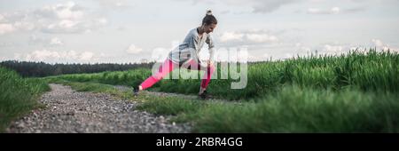 Junge Frau in pinkfarbenen Leggings, die sich streckt, bevor sie auf einem wunderschönen grünen Ackerland joggt. Weitwinkelbild. Stockfoto