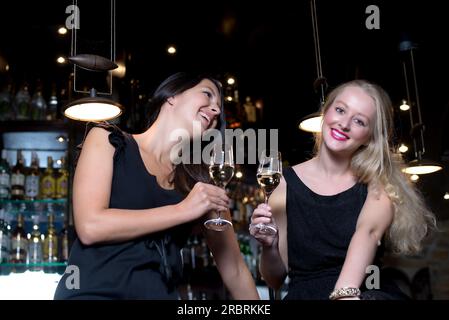 Zwei wunderschöne junge Frauen, die elegante schwarze Kleider tragen, feiern mit alkoholischen Getränken an einem schicken Ort Stockfoto