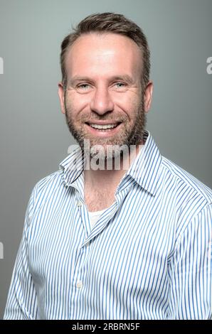 Porträt eines lächelnden, gut aussehenden Mannes mittleren Alters mit einem Bart, der ein gestreiftes Hemd trägt und in die Kamera schaut, mit einem warmen, freundlichen Lächeln gegen einen Stockfoto