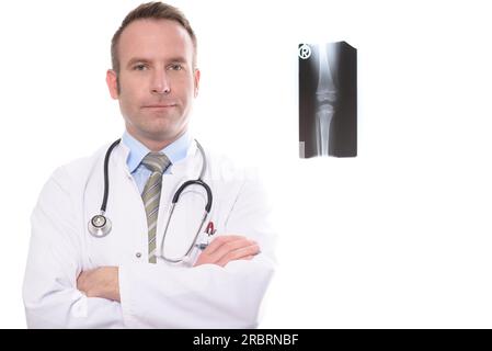 Selbstbewusster männlicher Arzt oder Arzt in einem Laborkittel und Stethoskop, der mit gefalteten Armen vor dem Röntgenbild eines Kniegelenkes steht und auf den schaut Stockfoto