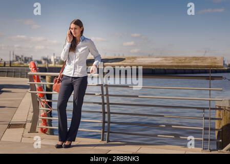 Eine Frau, die auf einem Mobilgerät steht, sich auf dem Geländer lehnt und an einem Pier oder einer Strandpromenade sitzt, während sie einen Sommertag an der Küste genießt Stockfoto