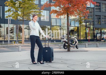 Aktive junge Geschäftsfrau, die mit ihrem Handy telefoniert, während sie draußen neben ihrem Trolley Case wartet und bereit für die Reise ist Stockfoto