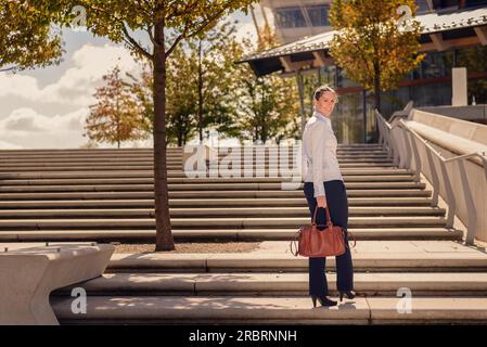 Stilvolle Frau in Hosen tragen einer Handtasche klettern einen Flug von konkreten Outdoor urban Treppen anhalten zu drehen und zur Seite schauen Stockfoto